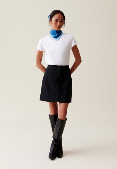 Camisa blanca de manga corta, falda negra de cintura alta con botones y bolsillos, combinada con botas negras hasta la rodilla y un accesorio de bufanda de seda azul.