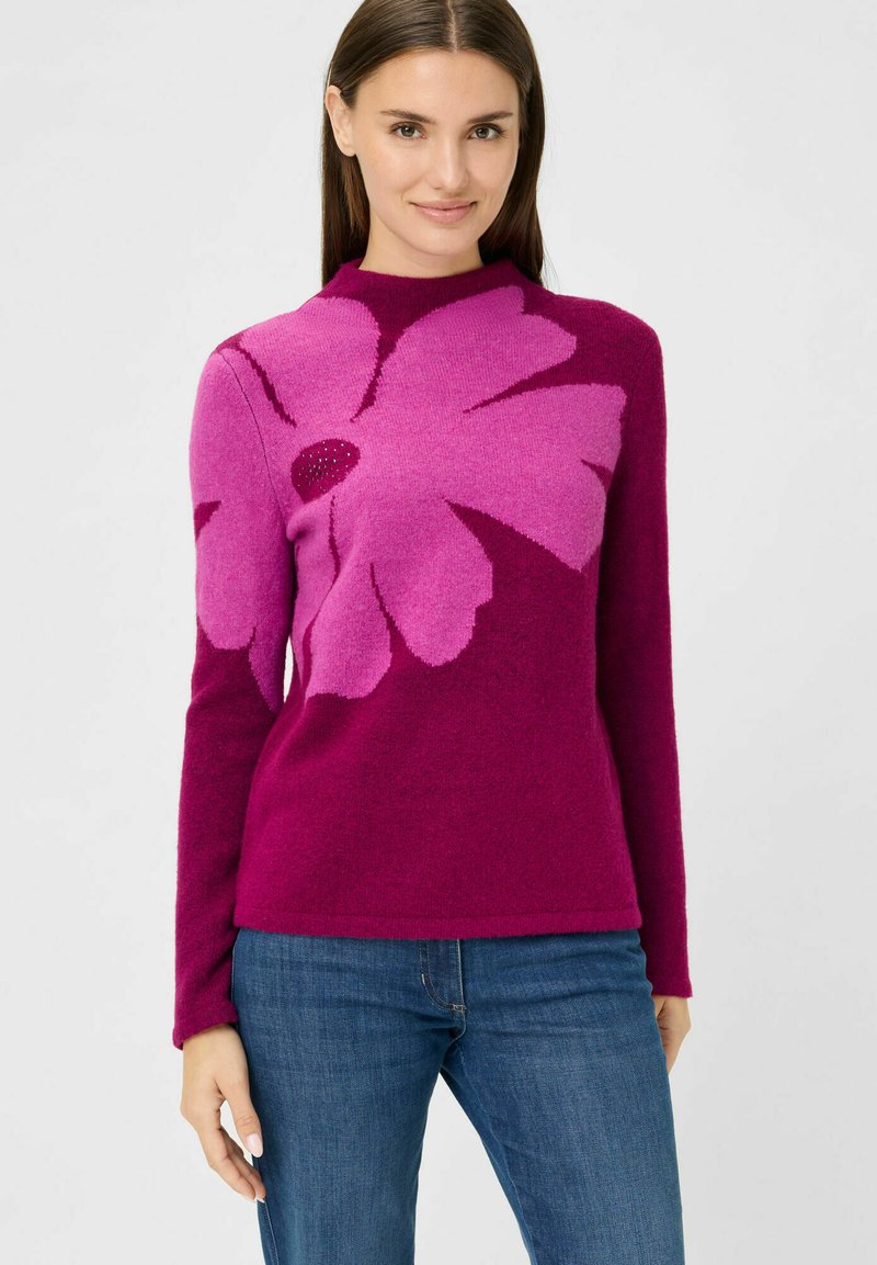 Fuchsia Pullover mit einem großen pinken Blumenmuster, hohem Kragen, langen Ärmeln und weichem Material, kombiniert mit blauen Jeans.