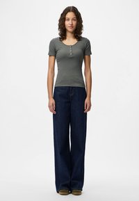 Top henley cinza com riscas e mangas curtas emparelhado com jeans de corte largo em denim escuro e ténis verdes-oliva. O sujeito está posicionado contra um fundo branco.