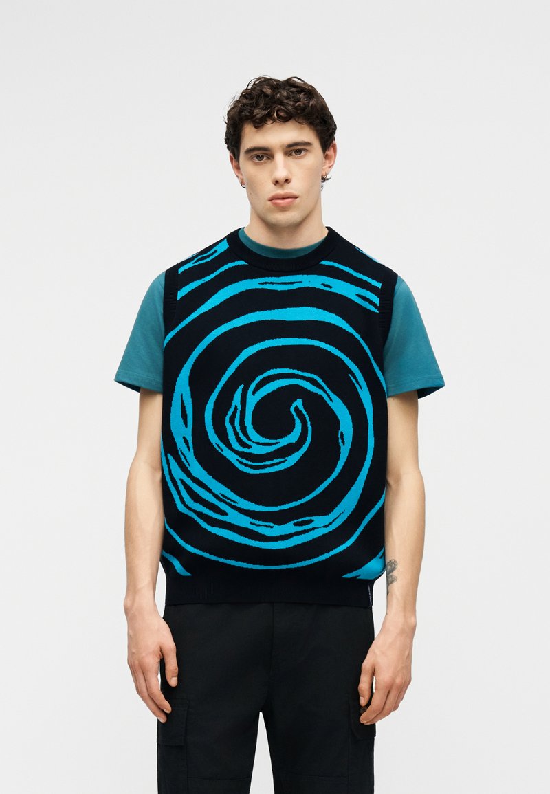 Νεαρός με κοντά σγουρά μαλλιά φοράει ένα μαύρο αμάνικο γιλέκο με μεγάλο μπλε σπειροειδές σχέδιο πάνω από μια τιρκουάζ κοντομάνικη μπλούζα.