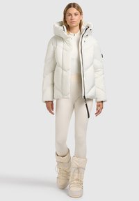 White puffer jacket with a hood, featuring a quilted design, zip closure, and black accents. Worn over a light-colored outfit and boots.