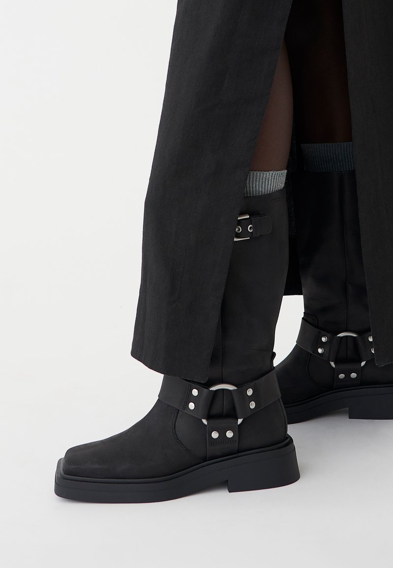 Schwarze Ankle Boots aus Leder mit mattem Finish, rechteckiger Zehenpartie und dekorativen Schnallen. Die Sohle ist klobig und strukturiert.