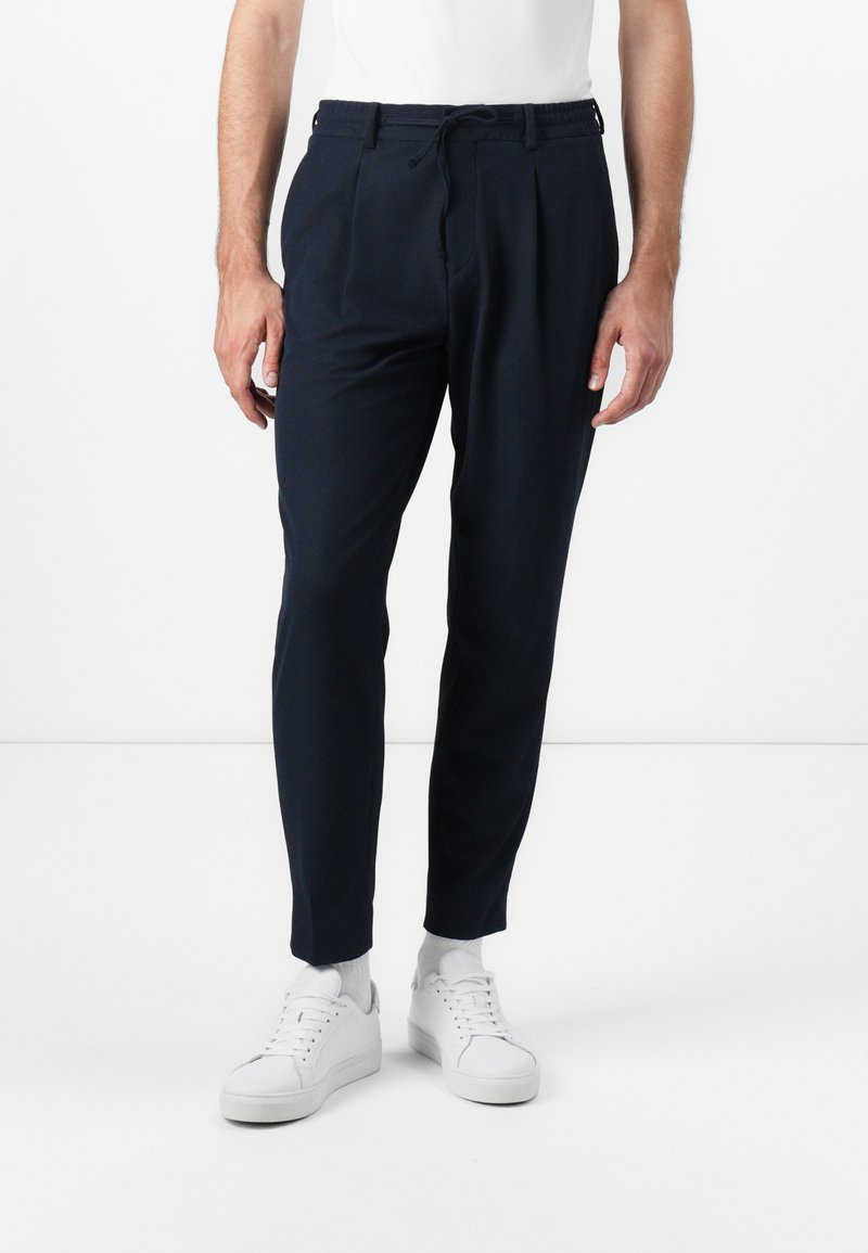 Navy tapered broeken met plooien, een trekkoord in de taille en zijzakken, gemaakt van een gladde stof. G gedragen met witte sneakers en sokken.