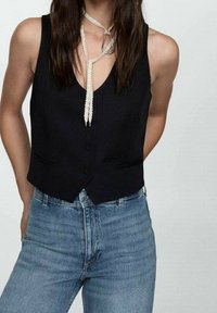 Chaleco negro sin mangas con diseño de escote en V, que incluye dos bolsillos delanteros y cierre de botones. Combinado con jeans azules de cintura alta.