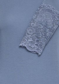 Světle modrý žebrovaný top s krajkovým detailem na rukávu, který obsahuje složité květinové vzory a vlnitý okraj. Celkově měkká textura.