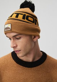 Brązowa dzianinowa czapka beanie z czarnym logo, z pomponem na górze. Tekstura jest prążkowana, z naszywką przedstawiającą szczegóły gór i słońca.