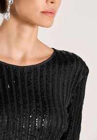 Frau in einem schwarzen, mit Pailletten besetzten Oberteil mit horizontalen Streifen und großen klaren Tropfohrringen, von Hals bis Brust in sanfter Beleuchtung gezeigt.