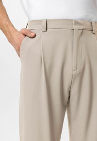 Beige broek gemaakt van gladde stof, met geplooide voorkant, zijzakken en een nauwsluitende pasvorm met een strak ontworpen tailleband.