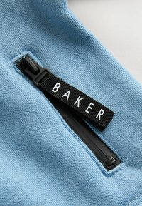 Tela de color azul claro con una cremallera y una etiqueta negra que dice "BAKER". La textura es suave y la cremallera está situada a lo largo de la costura.