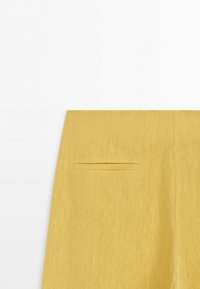 Close-up van een gele stoffen broek met een enkele achterste paspelzak, eenvoudige stiksels en een schone, gladde textuur tegen een witte achtergrond.