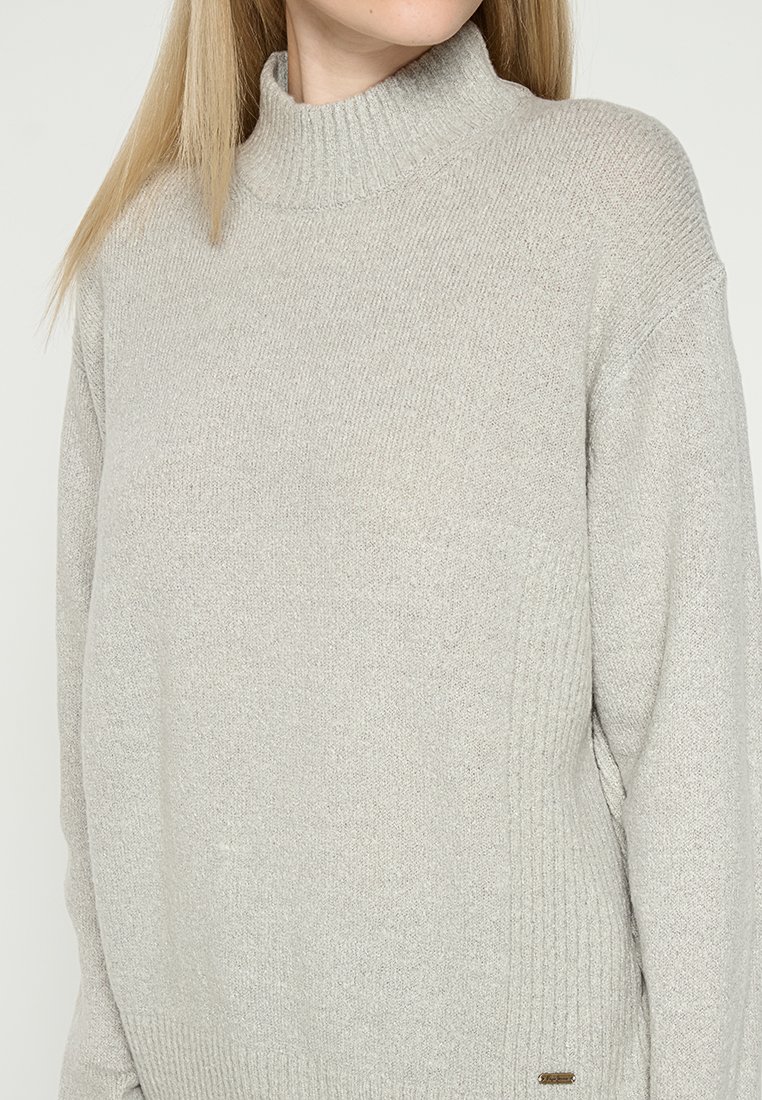 Maglione a dolcevita grigio chiaro realizzato in morbido tessuto a maglia. Presenta una texture a coste e una vestibilità ampia, con un piccolo tag metallico decorativo.