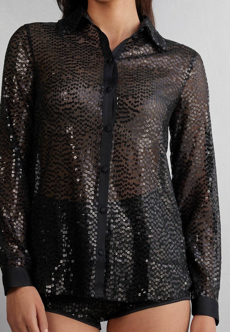 Femme portant une chemise noire transparente à manches longues avec des sequins noirs et argentés et un short noir assorti.