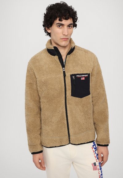 Tan fleece jacket with black zippered pocket, high collar, and black accents. Soft texture, zip closure, and contrasting branding details.