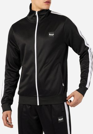 Mężczyzna ubrany w czarny dres z białymi paskami na rękawach i nogawkach, z zamykaną na zamek kurtką i spodniami oraz naszywkami z logo "Boxeur Des Rues".