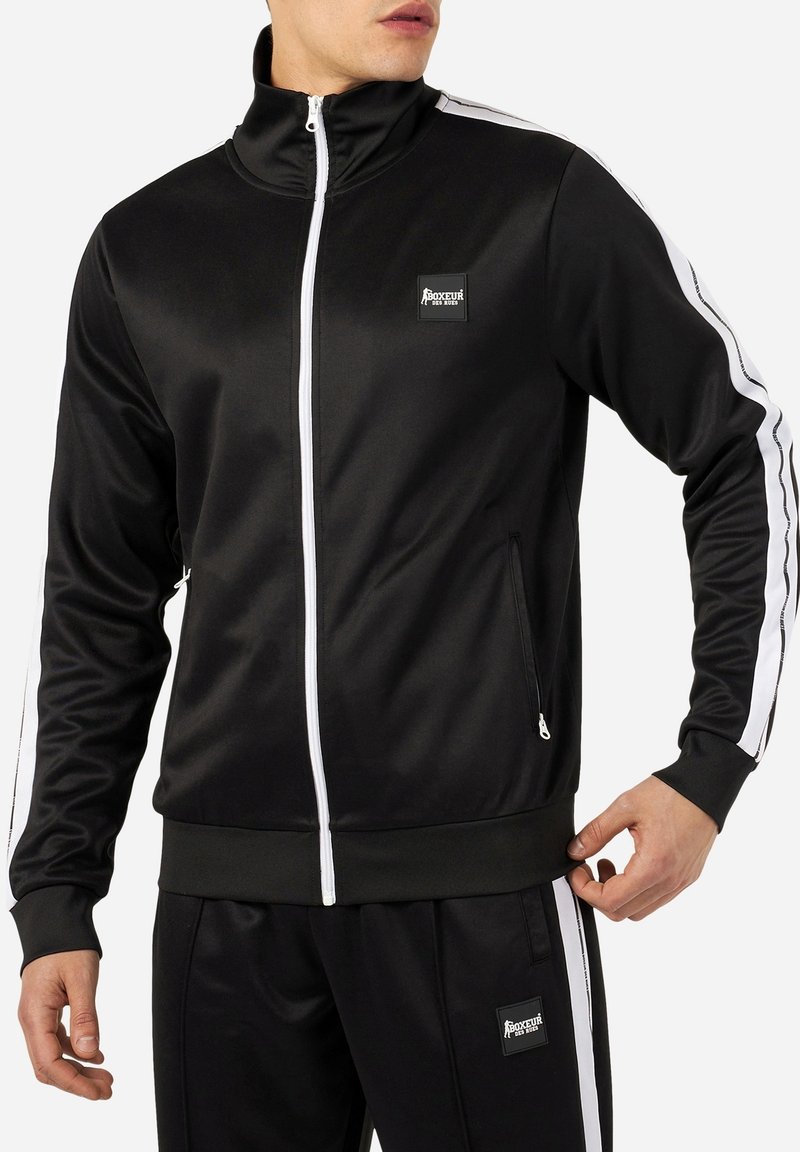 Homme portant un survêtement noir avec des bandes blanches sur les manches et les jambes, veste et pantalon zippés, avec des écussons logo "Boxeur Des Rues".