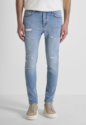 Hombre vistiendo jeans ajustados desgastados de color azul claro y zapatillas beige, de pie contra un fondo blanco liso.