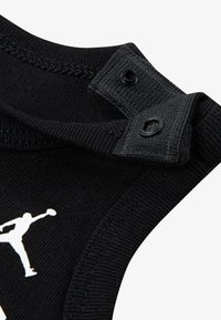 Mustast kangast rõivaese, millel on kaks musta pressnööpi, soonikuga kaelus ja valge hüppava figuuri graafiline logo.