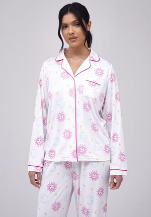 Woman wearing white pajama set with pink sun, blue moon, and gold star patterns, featuring pink piping and a chest pocket, standing against gray background.