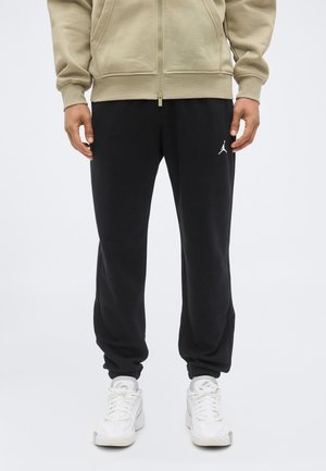 Mann trägt beige Zip-Up-Hoodie, schwarze Jogginghosen mit weißem Jumpman-Logo und weiße Sneakers, steht vor einem schlichten Hintergrund.