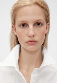 Vit skjorta med knappkrage, med en slät textur och krispig krage, bärs av en modell med rakt blont hår.