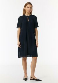 Marineblaues plissiertes Kleid mit Schlüssel-Ausschnitt und kurzen, schwingenden Ärmeln; der Stoff wirkt leicht und hat eine weiche Textur. Kombiniert mit schwarzen Schuhen.