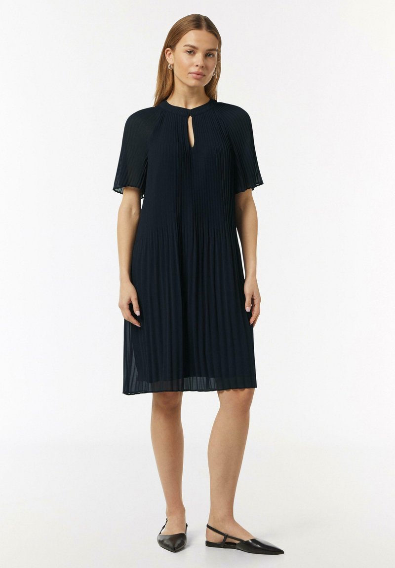 Marineblaues plissiertes Kleid mit Schlüssel-Ausschnitt und kurzen, schwingenden Ärmeln; der Stoff wirkt leicht und hat eine weiche Textur. Kombiniert mit schwarzen Schuhen.