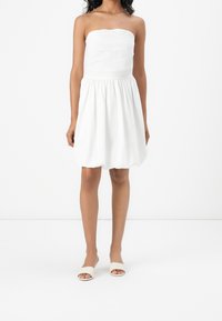 Bílé šaty bez ramínek s padnoucím živůtkem a nařasenou sukní. Hladká textura, délka ke kolenům, doplněné neutrálními sandály s otevřenou špičkou.