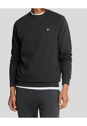 Man draagt zwarte sweatshirt met klein geel vogel logo op de linkerborst en bijpassende zwarte broek met subtiel logo op de linker dij.