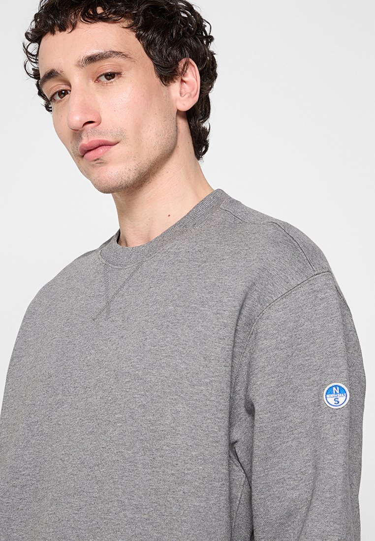 Hombre con cabello rizado y corto que lleva puesta una sudadera gris de cuello redondo sin estampado, con un pequeño logotipo azul circular en la manga.