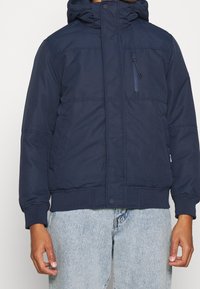 Chaqueta acolchada azul marino con capucha, que cuenta con un bolsillo con cremallera en la parte delantera y puños acanalados. Textura suave y diseño acolchado para aislamiento.