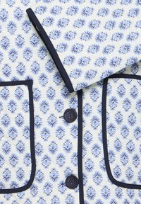 Material alb cu model floral albastru repetitiv, cu închideri cu nasturi negri și buzunare și guler cu margini negre.