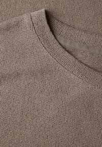 Hnědá pletená látka s texturovaným povrchem, obsahující žebrovaný kulatý výstřih a jemný povrch. Detailní záběr zdůrazňuje detaily materiálu.