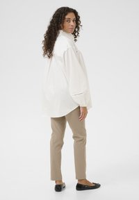 White, long-sleeve shirt with ruffled collar; beige pants; black flat shoes; curly hair. Shirt features loose fit and subtle detailing near cuffs.