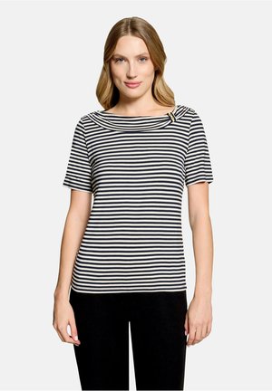 Femme portant un haut à manches courtes à rayures horizontales noires et blanches avec un col rond et un pantalon noir, debout devant un fond uni.