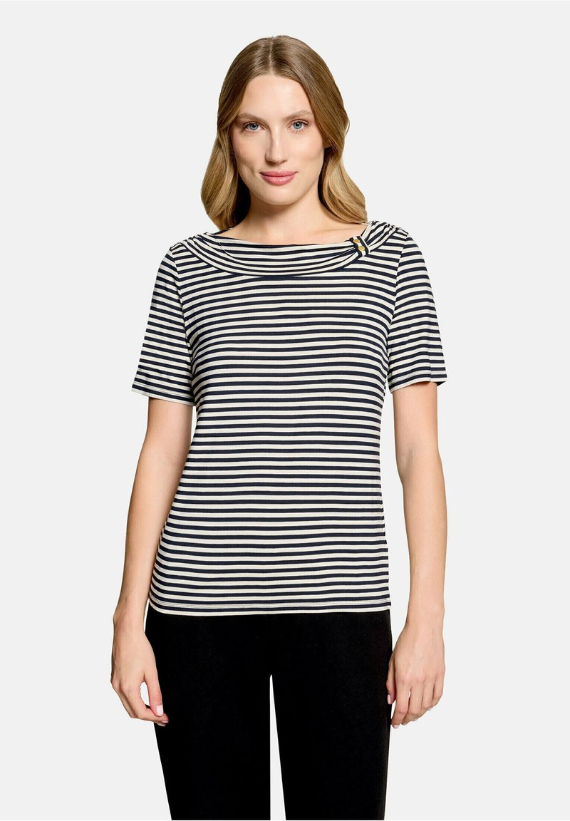 Femme portant un haut à manches courtes à rayures horizontales noires et blanches avec un col rond et un pantalon noir, debout devant un fond uni.