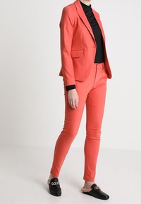 Korálový blazer a přiléhavé kalhoty s černým pleteným rolákem. Černé slip-on boty s řetízkovým akcentem. Hladká tkanina, módní střih.