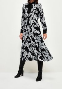 Vestido negro con intrincados patrones florales y de paisley en blanco, mangas largas y cuello con lazo. Llevado con botas negras hasta la rodilla.