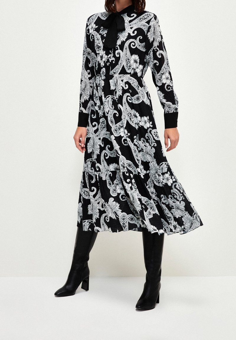 Vestido negro con intrincados patrones florales y de paisley en blanco, mangas largas y cuello con lazo. Llevado con botas negras hasta la rodilla.