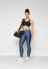 Svart sportsbh, marineblå leggings med hvit band, tettsittende design, glatt tekstur, og hvite joggesko. Modellen holder en svart treningsbag.