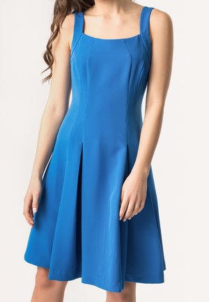 Kobieta w niebieskiej sukience bez rękawów sięgającej do kolan z szerokimi ramiączkami i plisami, stojąca na jednolitym białym tle.