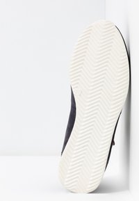 Shoe preto de camurça com uma sola de borracha branca texturizada com padrão em espinha de peixe. O perfil lateral mostra costuras e pequenos detalhes.