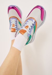 Multifarbige Sneakers mit weißen Leder, glänzenden lila, orangen und grünen Akzenten, einer strukturierten Gummisohle und weißen Stricksocken.