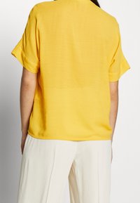 Top giallo a maniche corte con vestibilità rilassata, texture liscia e scollatura rotonda semplice, abbinato a pantaloni a gamba larga color crema.