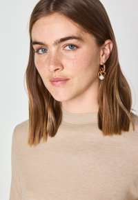 Top beige a collo alto realizzato in tessuto morbido. Presenta una superficie liscia e aderisce al corpo. Orecchini a cerchio dorati con un accento di perla.
