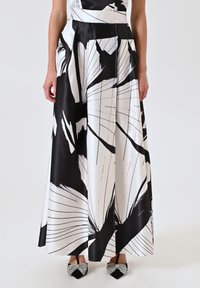 Jupe maxi imprimée florale en noir et blanc avec une finition satinée, présentant des plis et une taille ajustée, accompagnée de chaussures plates ornées de bijoux.