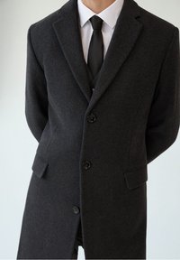 Donkergrijze wollen overjas met een gekleed kraag, twee voorzakken en drie zwarte knopen. Draag het over een licht shirt en een das.