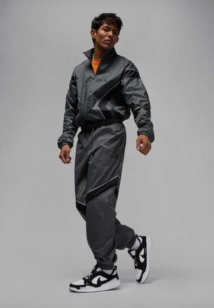 Joven con chándal deportivo gris y negro y zapatillas blancas y negras posando frente a un fondo liso de color gris claro.