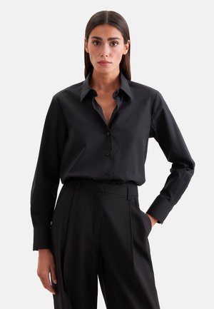 Vrouw met stijl donker haar, gekleed in een zwart overhemd met knopen en hoge zwarte broek, staand tegen een effen lichte achtergrond.