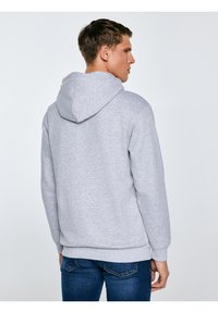 Noorest mehest vaate tagant, kes kannab kahvatuhalli kapuutsiga pusa ja siniseid teksapükse, taust on ühevärviline.