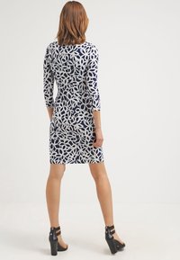 Navy dress with a white floral pattern, three-quarter sleeves, and a fitted silhouette. Black high-heeled sandals complete the outfit.